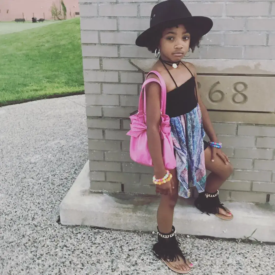 A young Leah Jeffries posing in a black hat, pink purse, and colorful skirt against a brick wall.